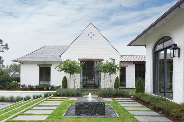 A landscaped courtyard view of a refined white-brick, single-storey residence. The composition features steep gabled roof forms, arched black-framed glazing, white masonry walls, a central rectangular water feature, and a formal garden with clipped topiary, ornamental grasses, lawn, and large rectangular stepping stones. The architecture has a traditional, symmetrical character with clean contemporary detailing.