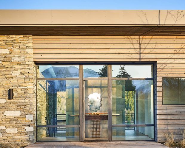 Exterior view of a contemporary, single-storey building with horizontal timber cladding, a stacked natural-stone wall, and a large dark-framed glazed entry or living space. Mountain scenery and trees are reflected in the glazing, while a sculptural interior feature is visible through the doors.