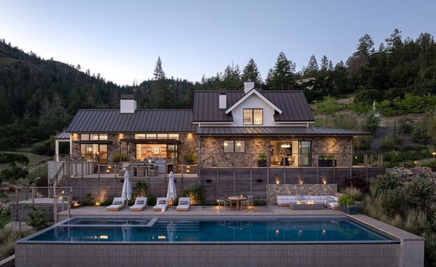 A dusk exterior view of a substantial mountain home with a dark metal gabled roof, stone-clad walls, extensive covered verandah, warm illuminated windows, and a large rectangular swimming pool in the foreground. The house is set into a wooded, sloping landscape and includes stepped terraces, outdoor seating, umbrellas, and landscaped planting.