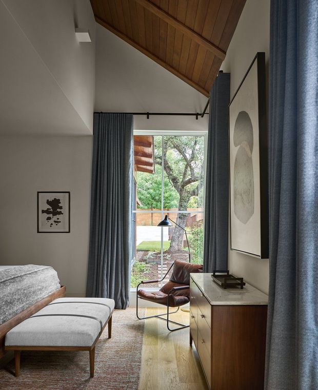 A warm, refined bedroom with a pitched ceiling clad in timber boards. The room features a low upholstered bed and matching bench, dark blue-grey full-height curtains, a mid-century leather lounge chair, timber dresser, framed artwork, and a large glazed opening overlooking a leafy outdoor area. Natural light, exposed timber, and neutral finishes create a calm, sophisticated atmosphere.