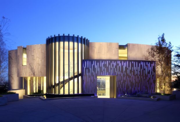 A dusk exterior view of a contemporary multi-volume building with warm timber-toned cladding, extensive floor-to-ceiling glazing, and a prominent curved glazed entrance volume framed by dark vertical fins. A perforated dark feature wall with narrow illuminated slots defines the front facade, opening onto a broad paved forecourt with low integrated planters.