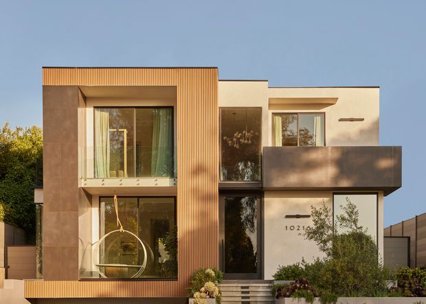 Front exterior view of a contemporary multi-level residence with a rectilinear composition, warm vertically ribbed timber-toned cladding, smooth light render, large floor-to-ceiling glazing, and projecting dark metal-clad balcony volumes. Landscaping and entry steps frame the facade.