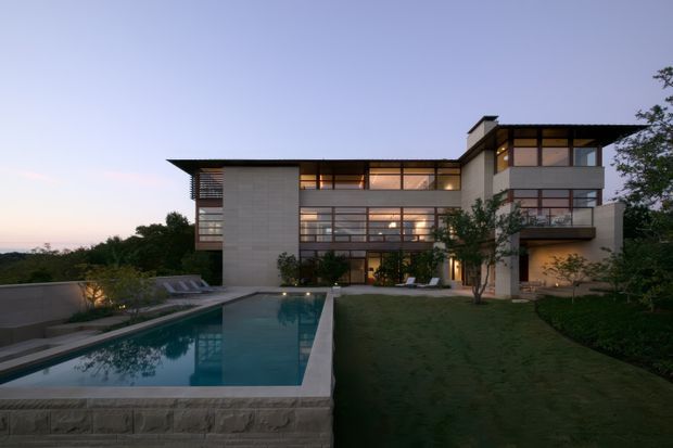 A contemporary multi-level residence at dusk, composed of rectilinear volumes with extensive floor-to-ceiling glazing, projecting balconies, and a broad low-pitched or flat roofline. The house overlooks a long rectangular swimming pool, with paved terraces, lawn, planted garden areas, and warm interior lighting visible through the windows.