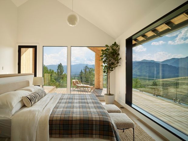 Bright contemporary bedroom with a low-profile upholstered bed dressed in layered white bedding and a muted plaid throw. Full-height glazing and a large corner window open to a timber deck with expansive mountain views. The room has white walls, a pitched ceiling, dark window frames, a simple spherical pendant, and a potted indoor tree, creating an airy alpine retreat atmosphere.