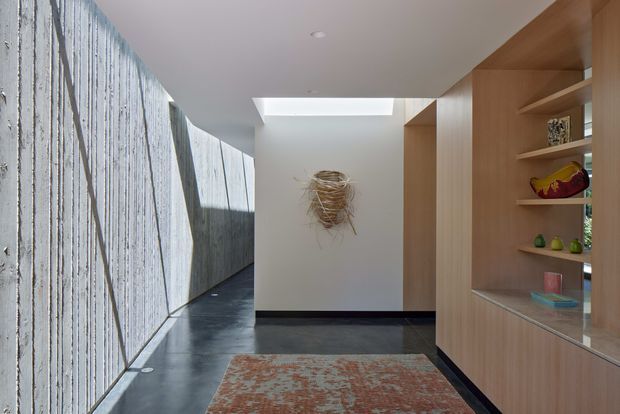 A refined modern hallway or entry passage with a long textured concrete feature wall and dark polished flooring on the left, contrasted by warm timber built-in shelving and display cabinetry on the right. A skylight brings natural light into the circulation space, with a patterned area rug and sculptural wall artwork adding warmth and visual interest.