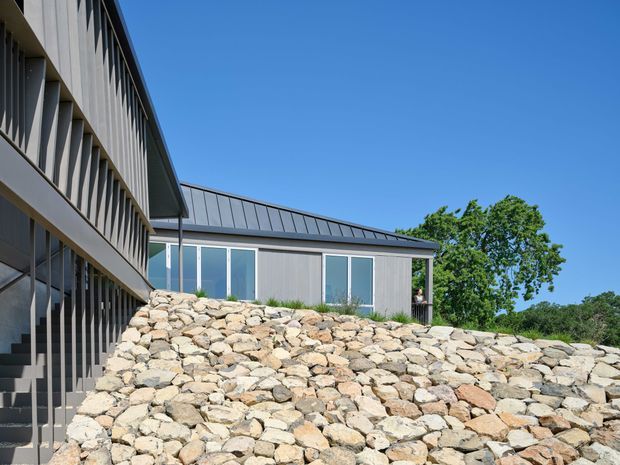 Exterior view of a contemporary multi-level building on a sloped site. The composition shows a dark grey metal-clad upper structure with strong vertical screening or fins, a lower pavilion with large glazed openings and a standing-seam metal roof, and a substantial natural stone retaining wall in the foreground. Clear blue sky and mature trees surround the building.
