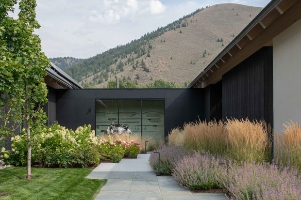 A modern single-storey residential courtyard entry framed by dark, vertically clad wings with pitched rooflines. A paved path leads through landscaped planting beds toward full-height glazed doors, with ornamental grasses and lavender-like shrubs in the foreground. Mountain terrain and woodland rise behind the building, creating a strong alpine setting. The composition is contemporary, restrained, and integrated with the landscape.