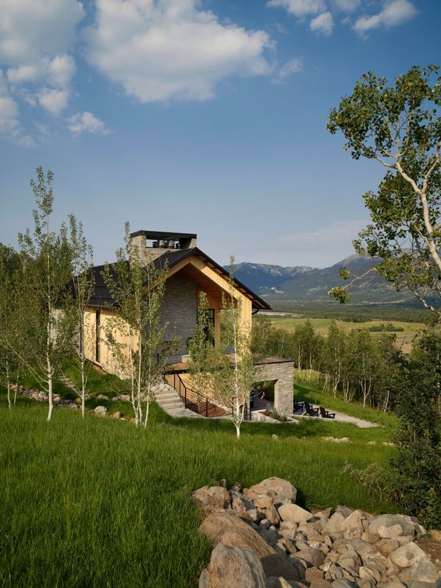 Exterior view of a contemporary mountain home set within a landscaped rural site. The house has a steeply pitched gable roof with dark roofing, stone and timber-toned exterior walls, large glazed openings, and a lower-level terrace or patio. A rocky drainage feature crosses the foreground, with lawn, young trees, open fields, and a mountain range beyond.