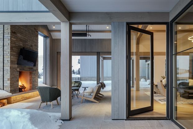 A refined contemporary living area opening onto a snowy outdoor setting. The interior features a substantial natural-stone fireplace wall with a mounted television, exposed structural timber posts and beams, grey vertical timber wall cladding, large black-framed glazed doors, and a neutral lounge arrangement. A secondary sitting area is visible beyond the central opening, while the right-hand glazing reveals another furnished interior space.