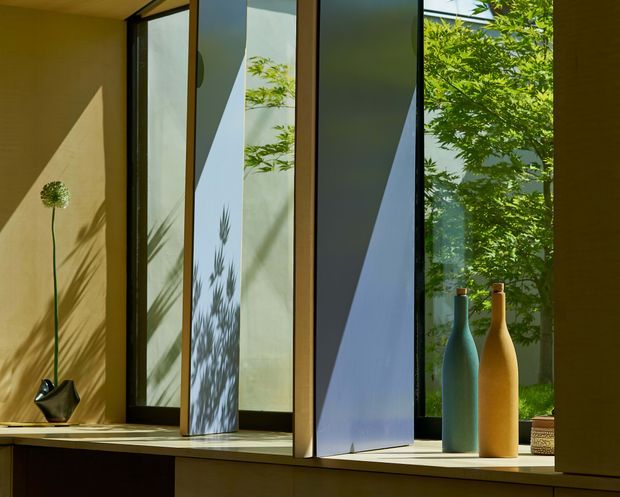 A close-up interior view of a sculptural window bay with tall, narrow floor-to-ceiling glazing and deep geometric mullions or fins. Strong sunlight creates angular shadows across warm ochre walls and the window reveal. A built-in timber-toned sill displays minimalist decorative vases and a small plant, with lush greenery visible outside. The image emphasizes daylight, architectural detailing, and the relationship between interior and garden.