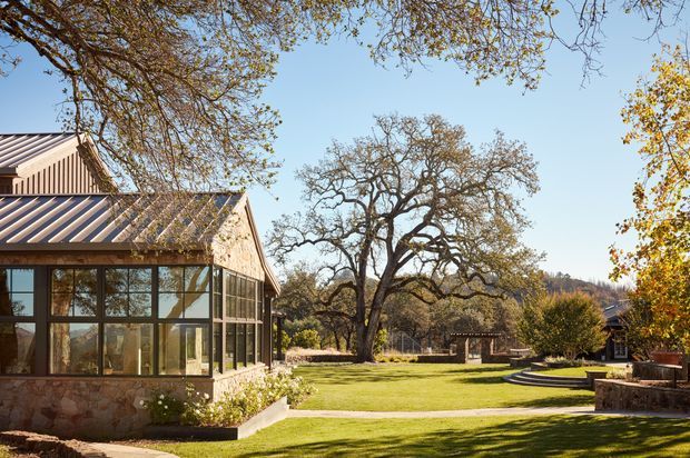 Exterior view of a low-rise residential building with a glazed sunroom or living extension, corrugated pitched metal roofing, vertical timber-like cladding, and stone masonry base walls. The building opens onto a landscaped lawn with mature trees, a paved path, raised stone-edged planting beds, and a small outdoor seating or patio area. Clear daylight and a rural, garden-oriented setting dominate the image.