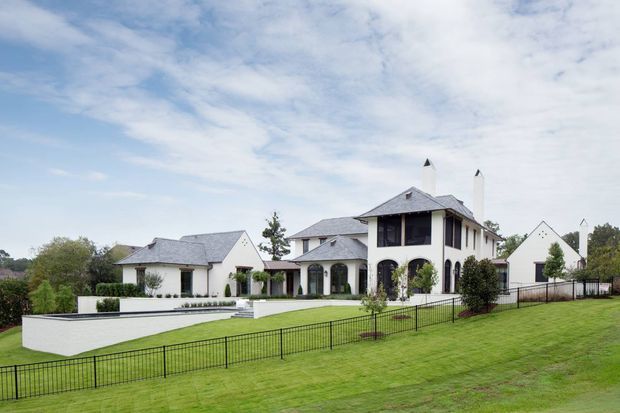 A large white multi-wing residential estate viewed from across a manicured lawn. The house features steep grey shingle roofs, multiple gables, tall chimneys, arched openings, dark-framed windows, covered verandas, and a formal rectangular pool or water feature in the foreground courtyard. Black metal fencing and landscaped planting define the property.