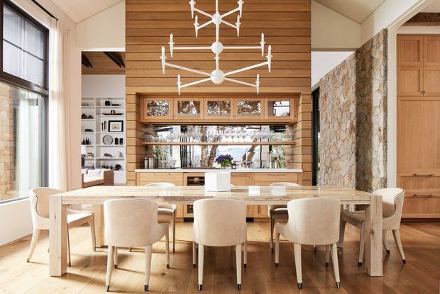 A bright, refined dining room with a large light-wood dining table surrounded by upholstered dining chairs. The room features a dramatic white sculptural chandelier, warm horizontal timber wall panelling, extensive built-in timber cabinetry and shelving, a large window, and a natural irregular-stone feature wall. The open-plan space appears connected to a living area and is illuminated by abundant daylight.