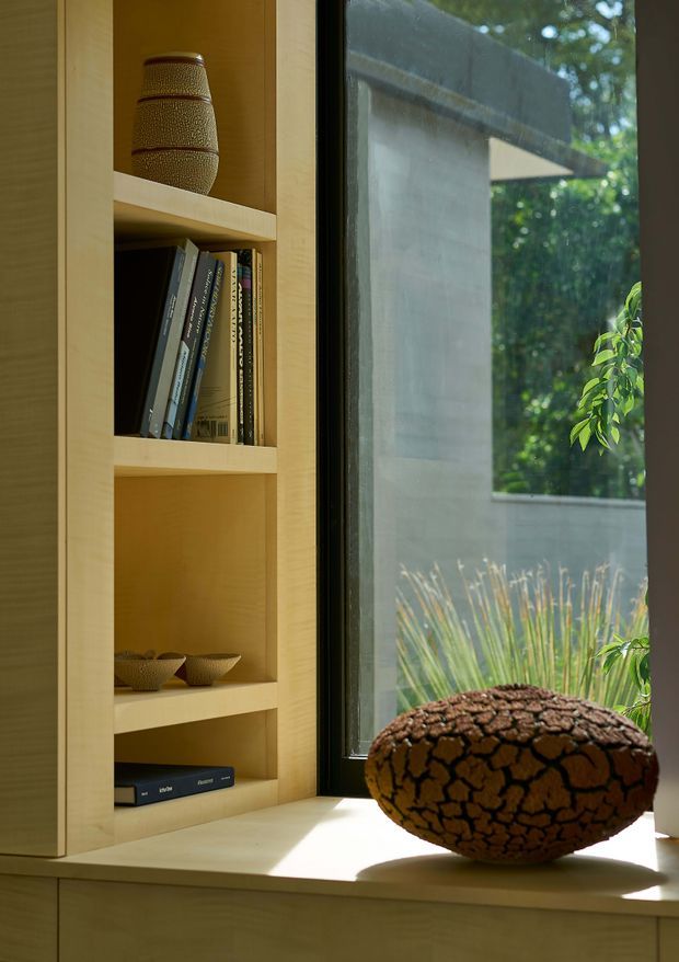 Close-up interior view of a built-in light timber shelving unit beside a large dark-framed window. The open shelves contain books, a ceramic vase, and small bowls, while a large textured decorative vessel sits on the deep timber window seat or sill. The window looks onto a leafy garden and an adjacent rendered wall. The composition is warm, refined, and natural, with strong emphasis on custom joinery and timber detailing.