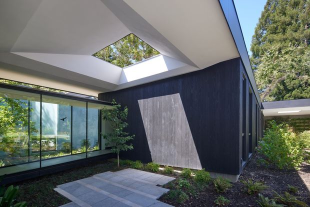 Contemporary courtyard view of a low-profile building with dark vertical cladding, extensive floor-to-ceiling glazing, a large geometric roof opening or skylight, and pale paved paths surrounded by established landscaping. The composition highlights the exterior architecture and connection between enclosed spaces and the courtyard.