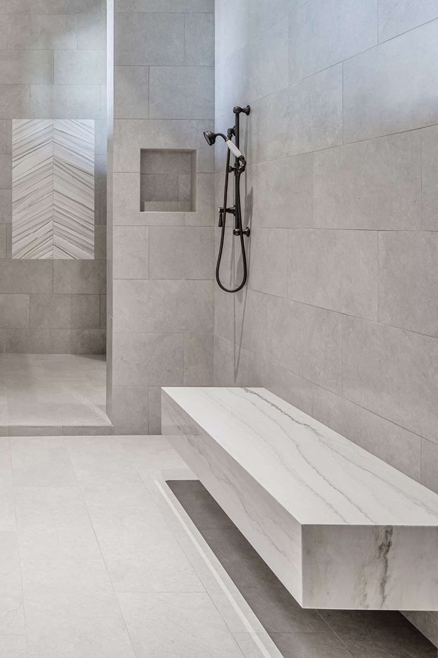 A minimalist contemporary bathroom featuring a large walk-in shower, pale grey stone-look wall and floor tiles, a recessed wall niche, dark metal shower hardware, and a substantial white marble-look floating bench or shelf with grey veining. The space is monochromatic, clean-lined, and softly lit.