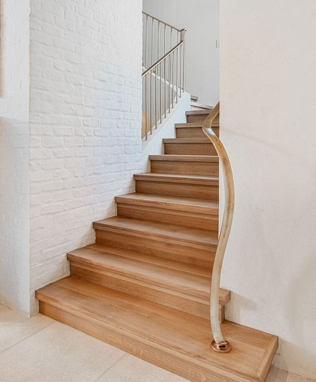 Bright interior photograph of a sculptural staircase. Broad timber-finished treads rise between white painted walls, with a white-painted exposed brick wall on the left. A curved timber handrail and slender metal balustrade create a distinctive contemporary stair detail.