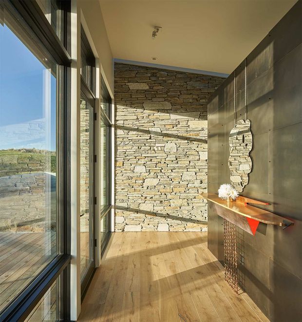 A narrow, sunlit interior passage or entry hall with full-height dark-framed glazing on the left, a rough stacked-stone feature wall at the end, and warm timber flooring. The right wall is clad in dark metal panels and includes a slim timber console, hanging decorative mirror, and small sculptural accessories. The glazing opens views to a stone terrace and landscape beyond. The composition combines rugged natural stone, timber, metal, and strong daylight in a refined contemporary setting.