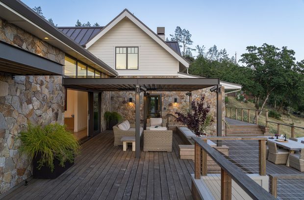 Exterior view of a multi-level residence with a broad timber deck and covered outdoor entertaining area. The facade combines extensive irregular natural stonework with light horizontal weatherboard cladding, dark metal roofing, timber decking, and glazed openings. Outdoor lounge and dining furniture occupy the deck, which steps down toward a landscaped sloping site with trees and a timber balustrade.
