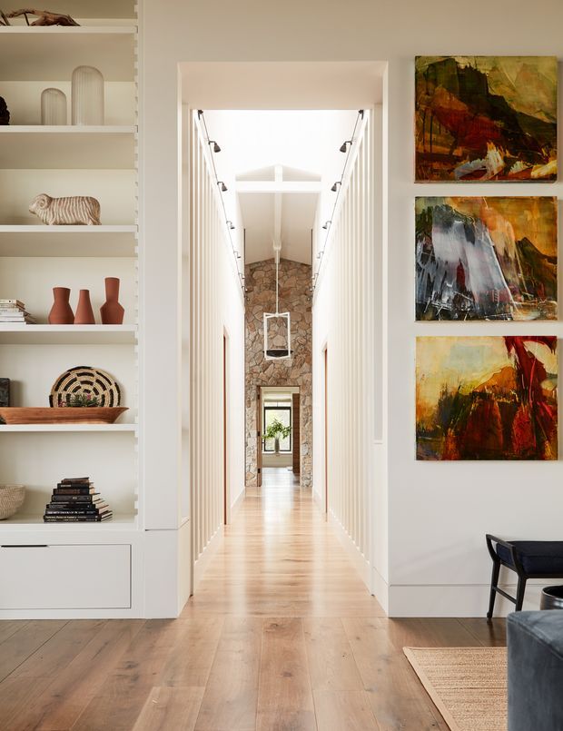 Bright, refined residential hallway viewed from a living area, with wide light timber flooring, white walls, built-in open shelving and cabinetry displaying ceramics and books, and three colourful abstract artworks. The long corridor has vertically detailed wall panelling, recessed linear lighting, a high ceiling with clerestory-like glazing, and a stone-clad feature wall at the far end. Partial seating and a rug are visible in the foreground.