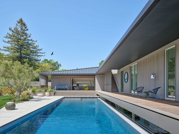 A contemporary single-storey residence viewed from the outdoor pool area. The long rectangular swimming pool is bordered by pale paving and a timber deck, with a covered outdoor living and dining area at the rear. The house has a low-pitched metal roof, extensive glazing, light vertical wall cladding, and a broad dark soffit. Landscaping and mature trees surround the property.
