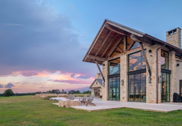 Exterior view of a large rustic-modern residence at dusk, set beside an expansive open lawn. The home features a dramatic exposed timber gable roof structure, pale stone masonry walls, extensive floor-to-ceiling glazing, dark-framed doors and windows, and a broad paved terrace with outdoor seating. A substantial stone chimney rises at the right, while the distant sunset landscape is visible beyond the property.