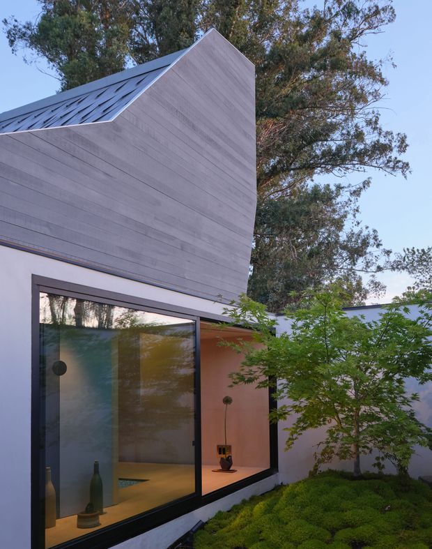Exterior view of a contemporary residence with a sculptural steeply folded roof form clad in pale grey timber-like panels, large dark-framed glazing, and a sheltered courtyard garden with a small ornamental tree and low groundcover. Warm interior lighting and simple decorative objects are visible through the windows.