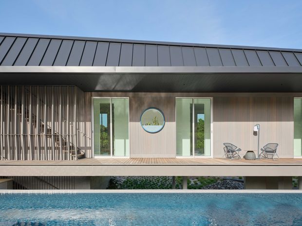 Contemporary multi-level residence viewed from across a swimming pool, featuring a dark standing-seam metal roof, vertically textured timber-toned facade, extensive glazing, a covered upper deck, and a screened outdoor area. Minimal outdoor furniture and the pool create a clean, resort-like composition.