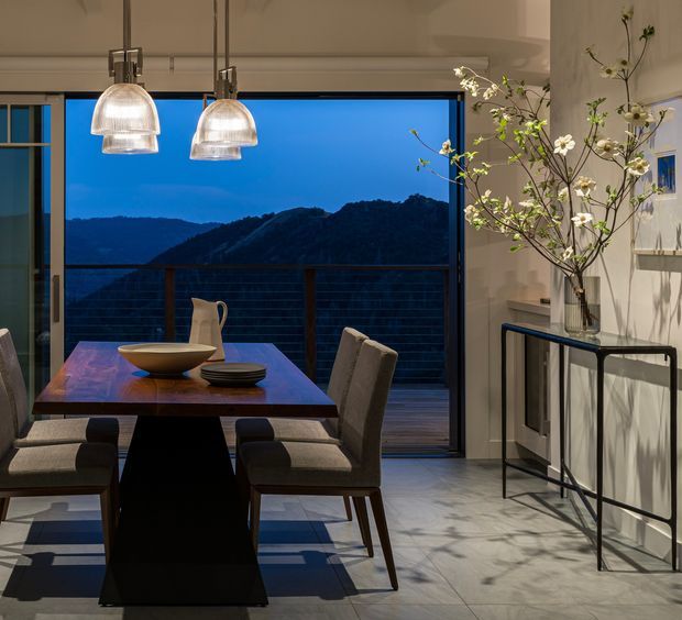 A refined dining area opening onto a balcony or terrace through large sliding glass doors, with expansive mountain views at dusk. The room features a dark wood dining table, upholstered dining chairs, contemporary pendant lights with ribbed glass shades, pale tiled flooring, and a slim black metal console topped with a tall flowering branch arrangement. The overall look is warm, modern, and sophisticated, with strong indoor-outdoor connection.