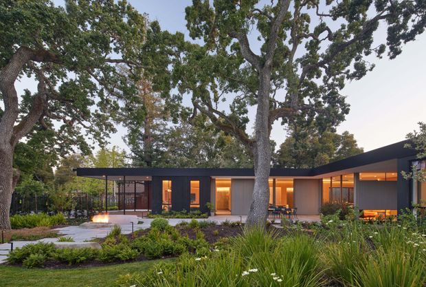 Single-storey modern residence set within a mature, landscaped garden. The dark, low-profile building has broad flat roof planes, extensive glazing, warm timber-toned interior illumination, and a sheltered outdoor entertaining area. Large established trees, ornamental grasses, layered planting, paved paths, and a small outdoor fire feature frame the facade.