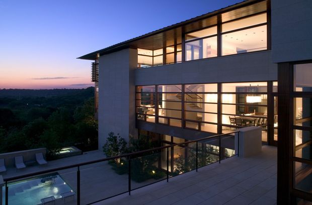 Twilight exterior view of a contemporary multi-storey house on a sloping, elevated site. The building has broad floor-to-ceiling glazing, dark slender window frames, pale masonry or rendered wall panels, projecting horizontal volumes, and warm interior lighting. A terrace with glass balustrades overlooks a wooded landscape, with a small illuminated pool or water feature visible below.