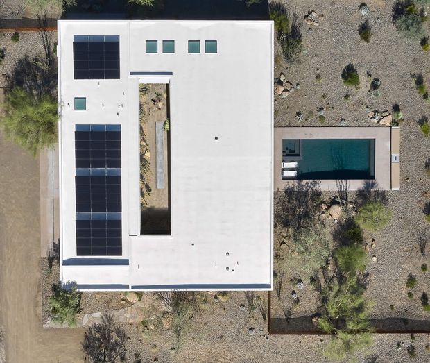 Overhead aerial view of a minimalist flat-roofed residence in a dry, rocky landscaped setting. The bright white rectangular roof contains multiple photovoltaic solar panel arrays and small skylights, with a narrow planted courtyard void through the centre. A separate rectangular swimming pool and paved deck sit alongside the house, surrounded by gravel, rocks, and sparse desert-style planting.