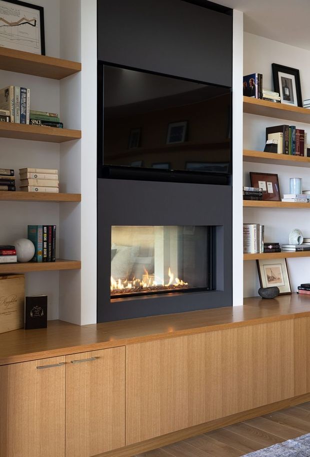 A built-in living room media and fireplace wall with a black inset television above a linear gas fireplace, framed by open timber display shelving. Below is extensive warm timber cabinetry with sleek horizontal handles. Books, framed artwork and small decorative objects occupy the shelves, creating a refined contemporary feature wall.