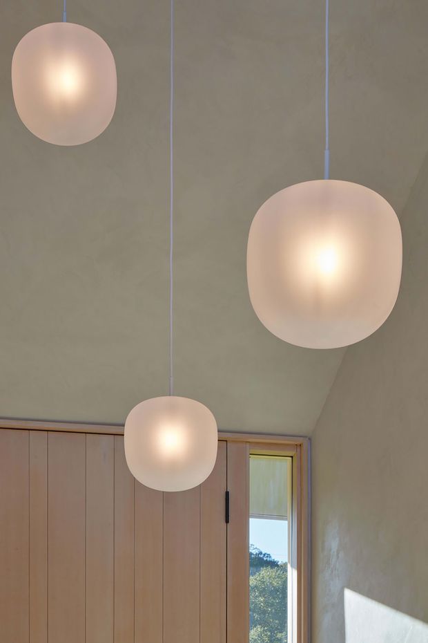 Interior detail showing three softly glowing, frosted globe pendant lights suspended at different heights from a high, sloping ceiling. The room has warm taupe plaster walls, vertical timber wall panelling, and a timber-framed window with sunlight entering from outside. The composition focuses on the lighting and ceiling volume, creating a calm, warm atmosphere.