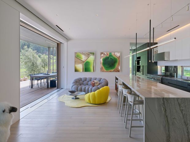 Bright open-plan contemporary living, dining and kitchen interior with pale timber flooring, extensive floor-to-ceiling glazing, and a large open connection to a timber deck and landscaped outlook. The living area features a sculptural grey rounded sofa, bold yellow lounge chair, abstract green artwork and a low coffee table. A long stone kitchen island with white bar stools occupies the right side, complemented by dark lower cabinetry, pale upper cabinetry, integrated appliances and slim linear pendant lighting. A close-up inset shows a veined grey stone finish, likely the island benchtop material.