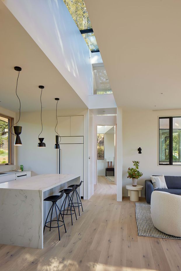 Bright open-plan interior combining a white marble-look kitchen island with black bar stools and pendant lights, pale timber flooring, and a partially visible living area with a blue sofa and textured rug. A central double-height void with clerestory-style glazing and a long skylight brings in abundant natural light. The room has clean contemporary detailing, white walls, concealed cabinetry, and black-framed windows.