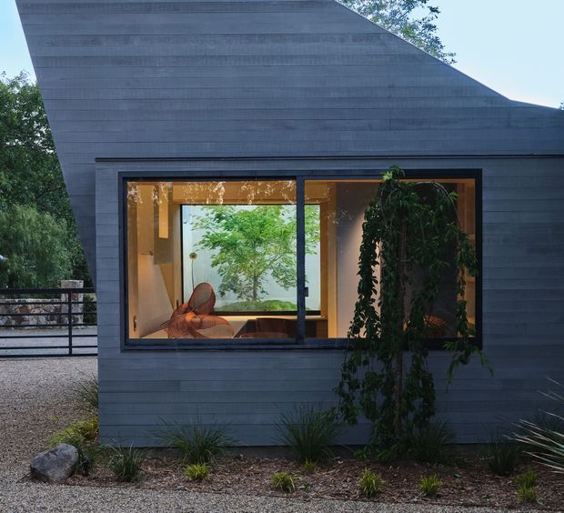 Exterior view of a compact contemporary building with dark grey horizontal cladding and a sharply sloping roof plane. A large black-framed window reveals a warmly lit interior with timber-toned surfaces, seating, and indoor planting. Gravel landscaping, grasses, rocks, and mature trees surround the structure.