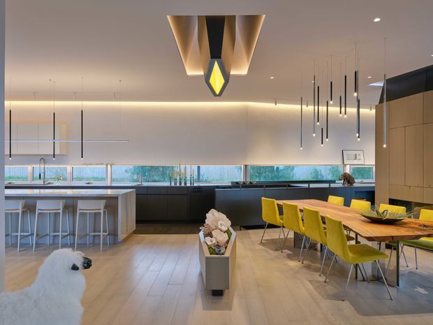 A spacious contemporary open-plan kitchen and dining area with a large island, integrated dark cabinetry, broad horizontal windows, and a long timber dining table surrounded by bright yellow chairs. The room features layered recessed and pendant lighting, light timber-look flooring, pale walls, and a sculptural central pendant. A small dog and decorative planter are present in the foreground.