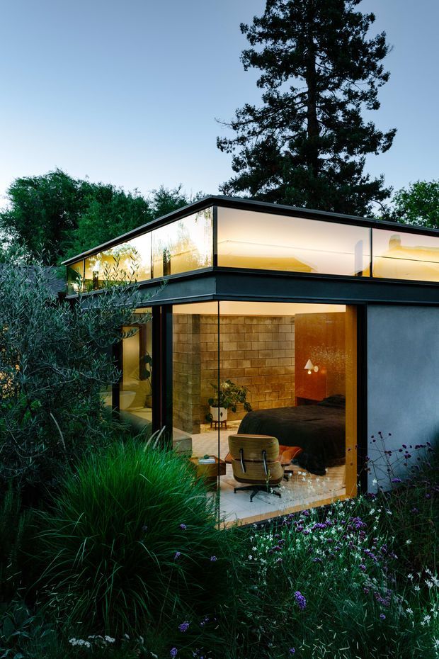 Twilight exterior view of a compact contemporary pavilion-like house set within dense landscaped planting. The building has a flat roof, crisp dark structural frames, extensive floor-to-ceiling glazing, and warm interior illumination. Through the glass, a bedroom with a dark bed, desk chair, and exposed block wall is visible. The surrounding garden includes ornamental grasses and flowering groundcover, while a tall mature tree rises behind the structure.