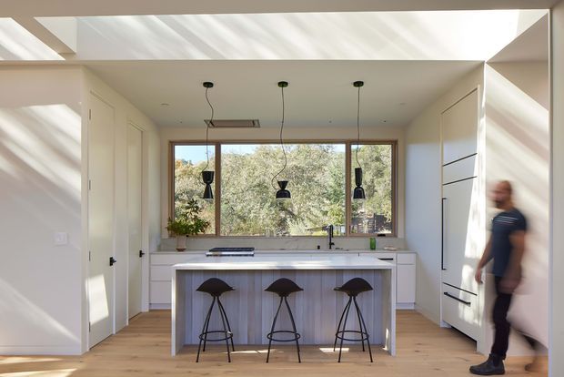 Bright contemporary kitchen with a central island and three bar stools, white built-in cabinetry, timber flooring, pendant lights, and a large timber-framed window overlooking trees. Strong natural light enters through a high-level skylight or clerestory opening, creating a clean, airy atmosphere.
