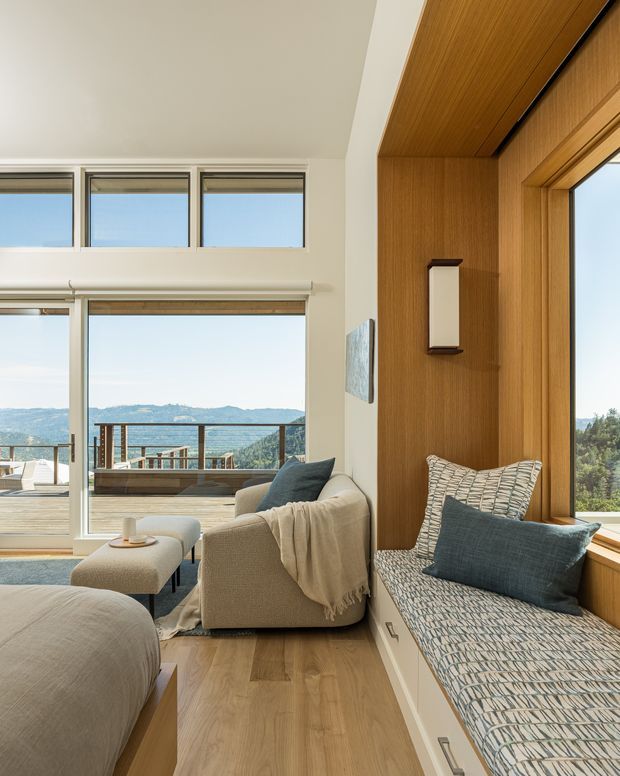 A bright contemporary living room with expansive glazing overlooking a mountainous landscape. The room features a built-in window seat with patterned upholstery and blue cushion, a neutral upholstered sofa and ottoman, timber wall and ceiling panelling, and a warm timber floor. A balcony or terrace is visible beyond the glazing.