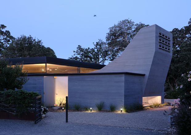 A modern single-storey residence photographed at dusk, with a strongly sculptural sloping concrete or rendered chimney-like volume, flat rooflines, extensive warm-lit glazing, and a minimalist landscaped gravel forecourt. Dark framing and horizontal wall planes contrast with the pale grey sculptural form, while mature trees surround the building.