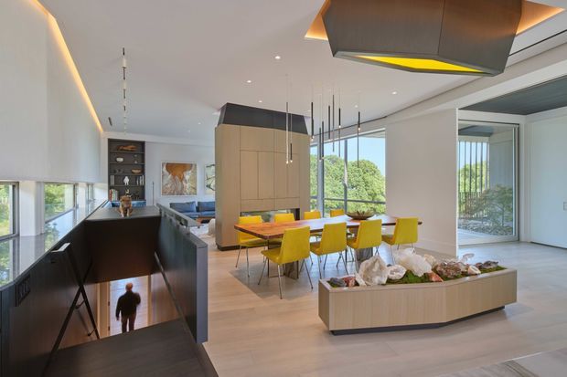Open-plan contemporary interior combining a dining area, living space and stairwell. A long timber dining table with bright yellow upholstered chairs sits beneath a sculptural suspended rangehood or ceiling feature, while a large timber-clad built-in volume separates the dining zone from the living area. Floor-to-ceiling glazing provides views to greenery, and a dark stair enclosure contrasts with pale timber flooring and white walls.