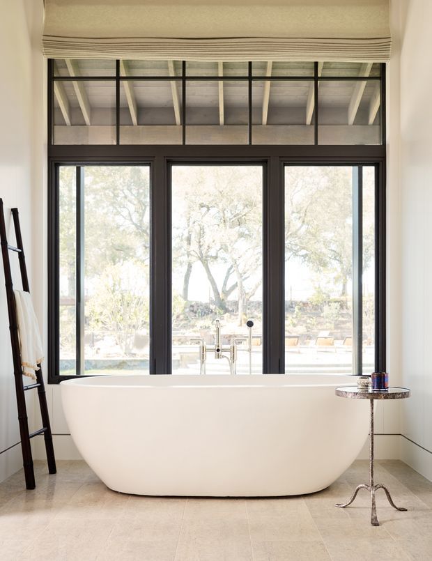 Bright, refined bathroom featuring a freestanding white soaking bathtub positioned in front of large black-framed windows. A high transom window incorporates exposed ceiling beams, while a Roman blind, pale tiled floor, chrome bath fittings, and a small round side table complete the space. Trees and an outdoor pool or patio are visible through the glazing.