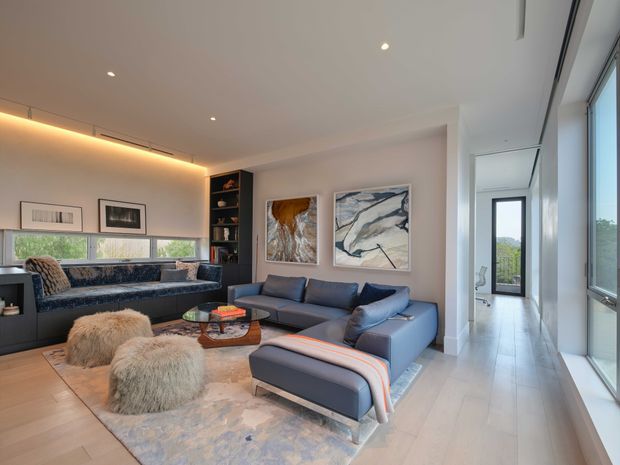 Contemporary living room with a large blue-grey sectional sofa, matching ottoman, built-in dark shelving, abstract artwork, and a low glass coffee table. Warm concealed cove lighting and recessed ceiling lights complement pale timber flooring, a soft area rug, and expansive glazing along the right side. A bright hallway or circulation space is visible beyond the living area.