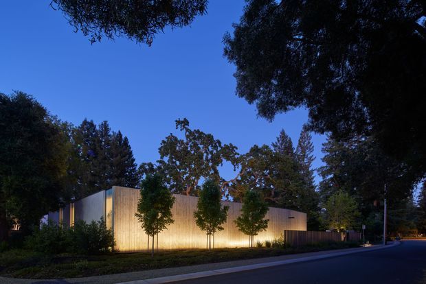 A contemporary single-storey building viewed from a quiet suburban street at dusk. The structure has a simple low-pitched or flat-roofed form with warm vertical timber cladding, pale rendered or panelled wall surfaces, and carefully integrated linear lighting washing the facade. Mature trees and landscaped planting frame the building, while the dark street and deep blue evening sky create a calm, dramatic setting.