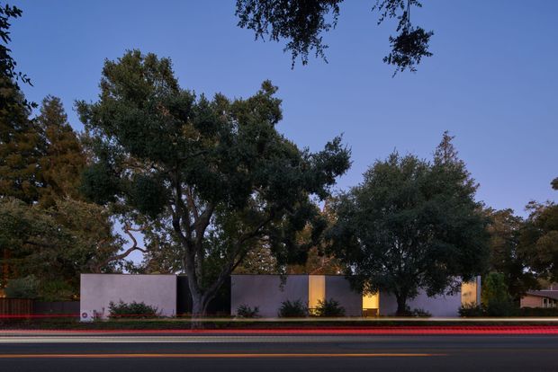 A low, modern single-storey building viewed from across a roadway at dusk. The restrained facade consists of pale rectangular wall planes with warm illuminated vertical openings, partly screened by mature trees and landscaping. Long-exposure light trails from passing traffic run across the foreground, creating a dramatic contrast with the quiet, horizontal architecture.