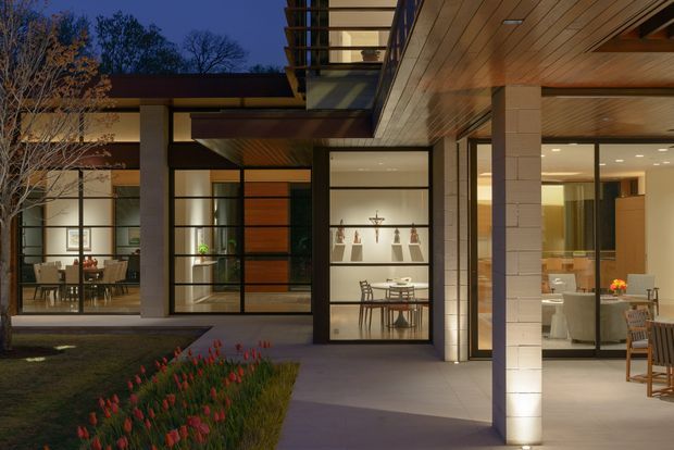 Twilight view of a refined modern residence organized around a landscaped courtyard. The composition highlights broad floor-to-ceiling glazing, dark metal-framed windows, warm timber soffits and cladding, pale masonry walls, and clearly visible dining and kitchen/living areas inside. Integrated lighting creates a warm, welcoming atmosphere.