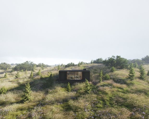 A small, dark, box-like architectural structure sits isolated in a broad natural grassland landscape. The compact building has a flat or very low-pitched roof, dark exterior cladding, and a large glazed front opening. Surrounding scrub, grasses, and low vegetation dominate the scene under a bright overcast sky.