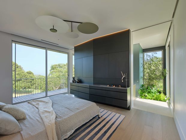 Bright contemporary bedroom with a low-profile bed, pale textured bedding, light timber flooring, and a large dark built-in wardrobe or media joinery wall. Floor-to-ceiling sliding glazing opens to a balcony with leafy views, while a glazed internal opening reveals a planted bathroom or lightwell. Minimal circular ceiling lights and warm concealed lighting add a refined modern feel.